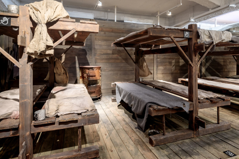 Sala wystawowa zaaranżowana na salę więzienną, w której spali zesłańcy na Sybir. Na pierwszym planie proste, drewniane prycze zaścielone brudnymi, cienkimi, postrzępionymi kocami. 
