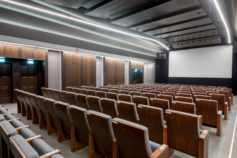 Sala kinowa utrzymana w kolorach szarości i brązu. Na pierwszy planie znajdują się rzędy drewnianych foteli kinowych o szarym obiciu. W głębi duży, biały ekran kinowy. 